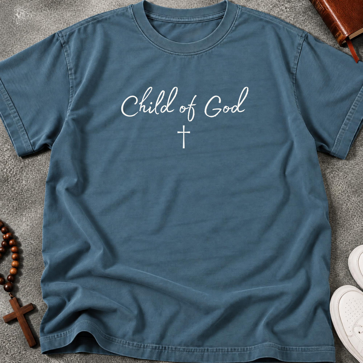 A slate blue crewneck t-shirt with 'Child of God' and a cross printed in white on the chest, laid flat on a gray textured surface with a wooden cross, rosary, and shoes as props.