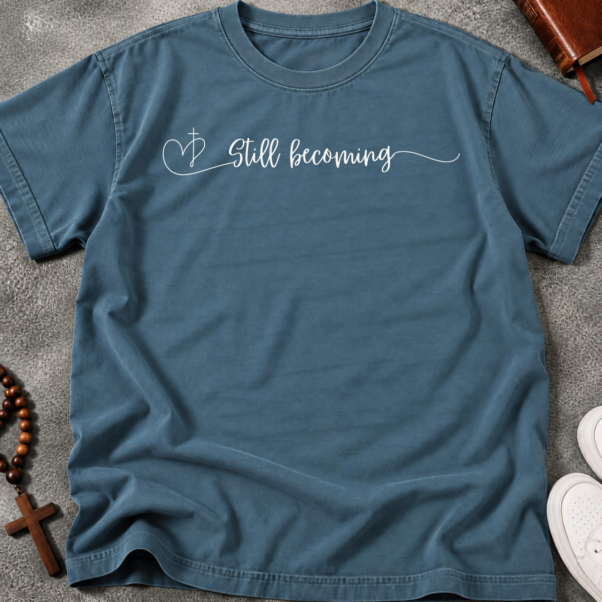 A steel blue short-sleeve t-shirt with 'Still becoming' written in white script across the chest, styled in a flat lay on white wood planks with blue jeans, a book, and a wooden bead bracelet nearby.