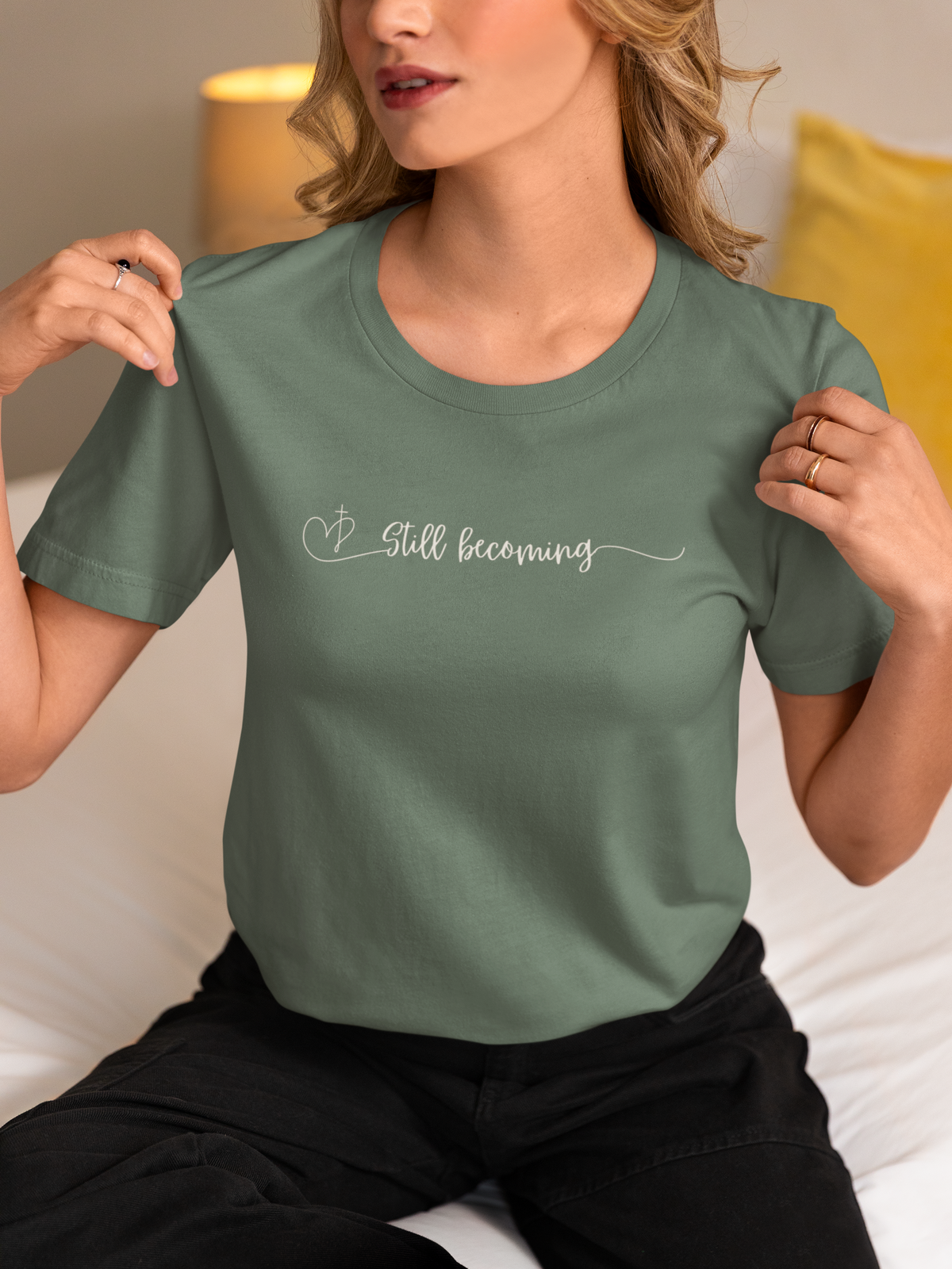 A woman wearing a sage green crewneck Christianity t-shirt by SeedSowerGraphics with the words 'Still becoming' written in white script across the chest, paired with black pants, sitting in a cozy bedroom setting.