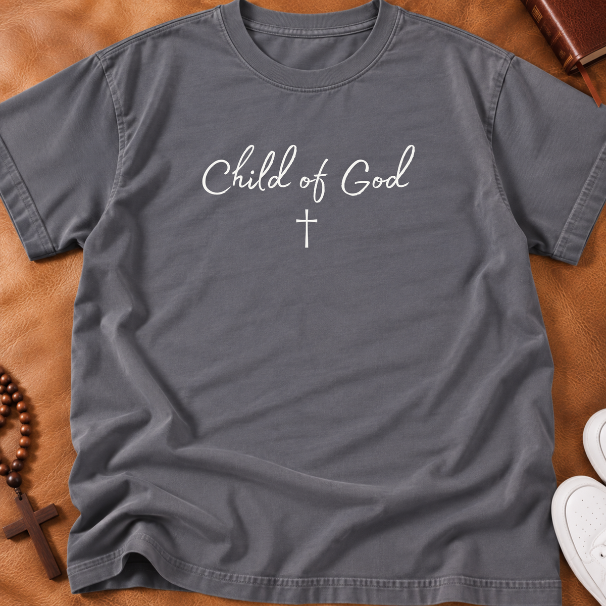 A dark gray crewneck t-shirt with 'Child of God' and a cross printed in white on the chest, laid flat on a brown leather background with lifestyle props.