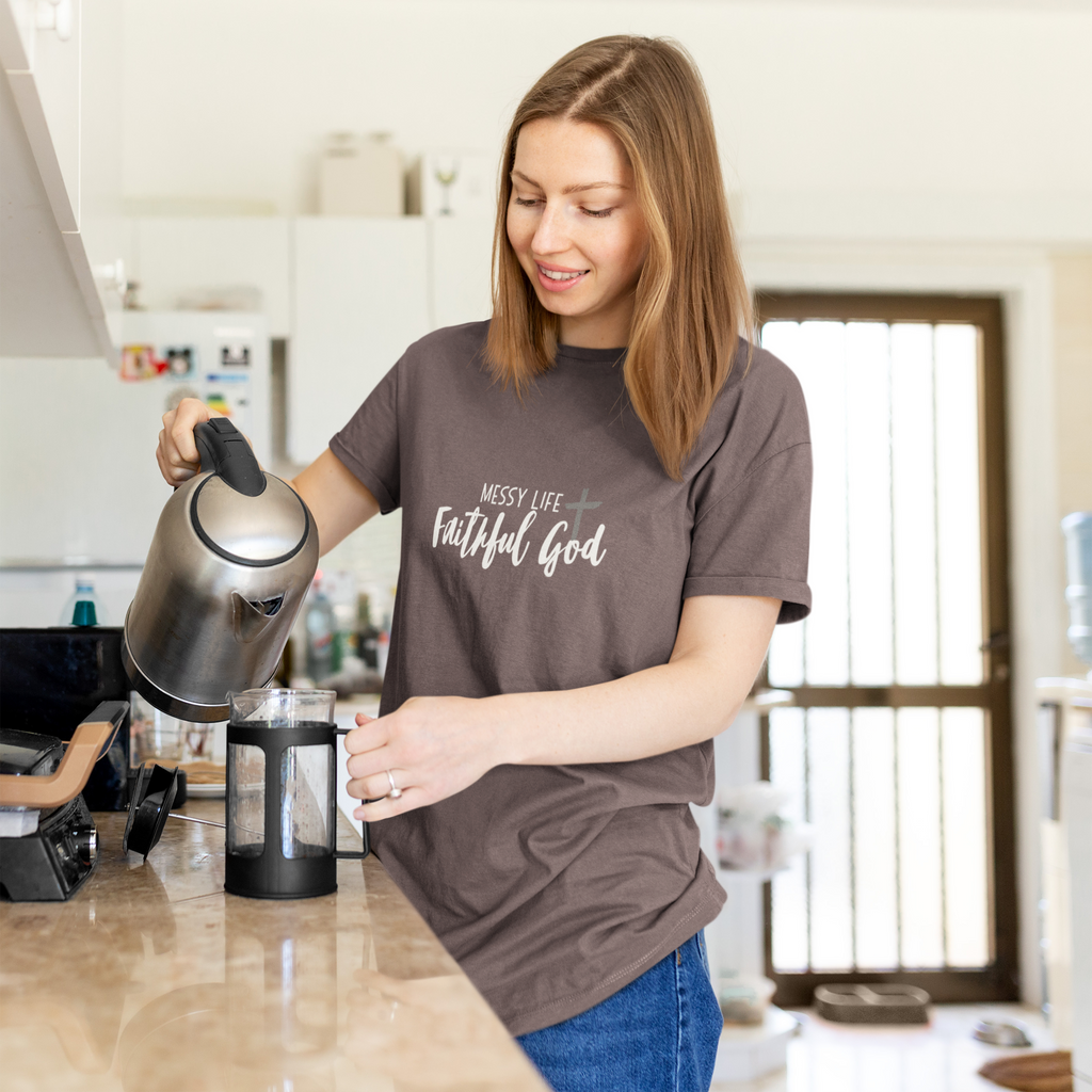 A woman in a kitchen wearing a brown Christian graphic t-shirt by SeedSowerGraphics  that reads 'MESSY LIFE Faithful God' in white script. She is pouring hot water from a stainless steel kettle into a French press.