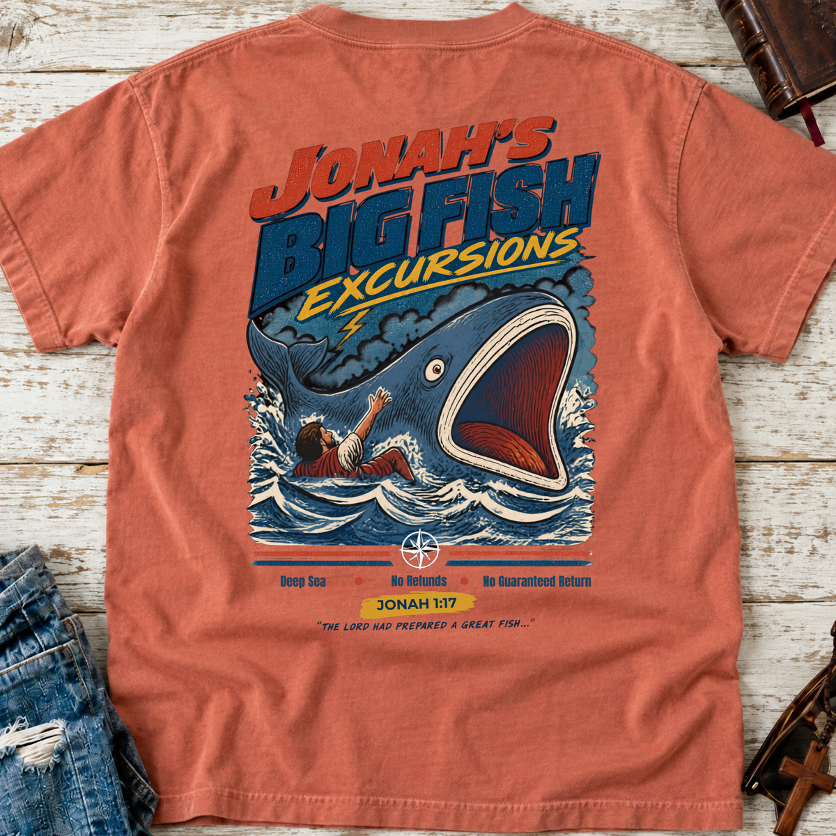 A coral orange graphic t-shirt laid flat on a whitewashed wooden surface, styled with a pair of distressed blue jeans. The back of the t-shirt features a large colorful graphic of a giant blue fish jumping out of the water with a man reaching up, under the text 'JONAH'S BIG FISH EXCURSIONS'.