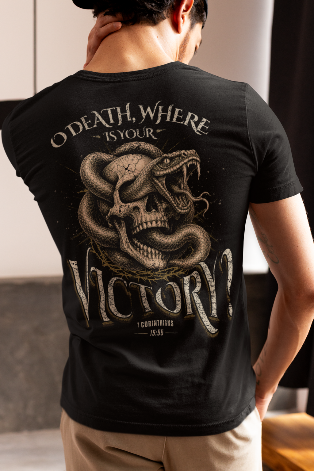 A man shown from behind wearing a black Christian graphic t-shirt. The graphic features a coiled snake wrapped around a human skull, with the text 'O DEATH, WHERE IS YOUR VICTORY?' in a distressed, vintage-style font.