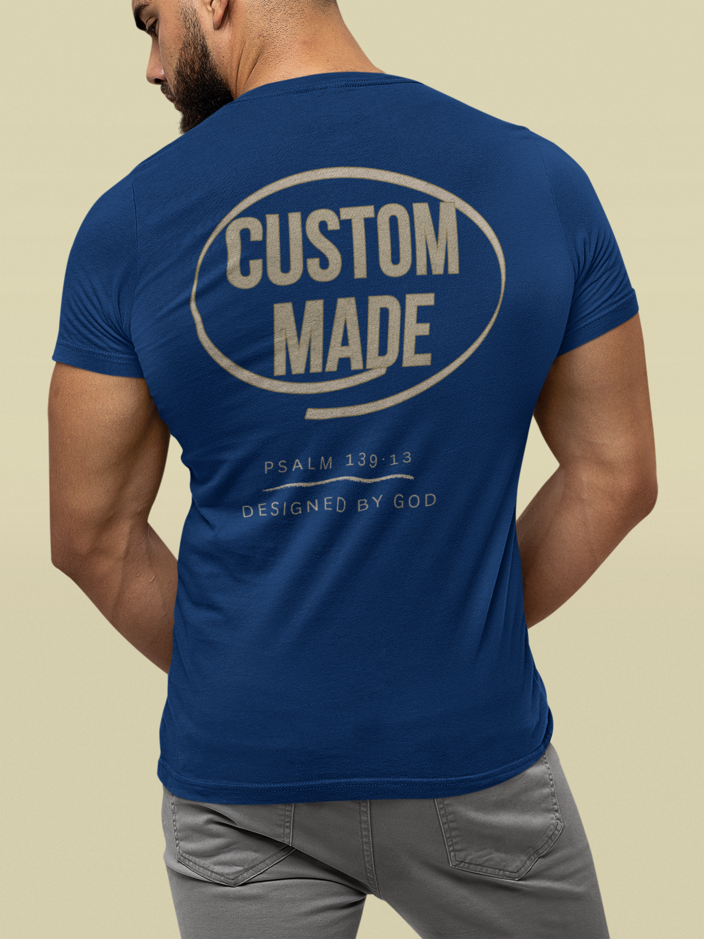 A man shown from the back wearing a royal blue Christian graphic t-shirt with a large cream-colored graphic on the back that reads 'CUSTOM MADE' inside an oval, with smaller text below. The background is a solid light beige color.