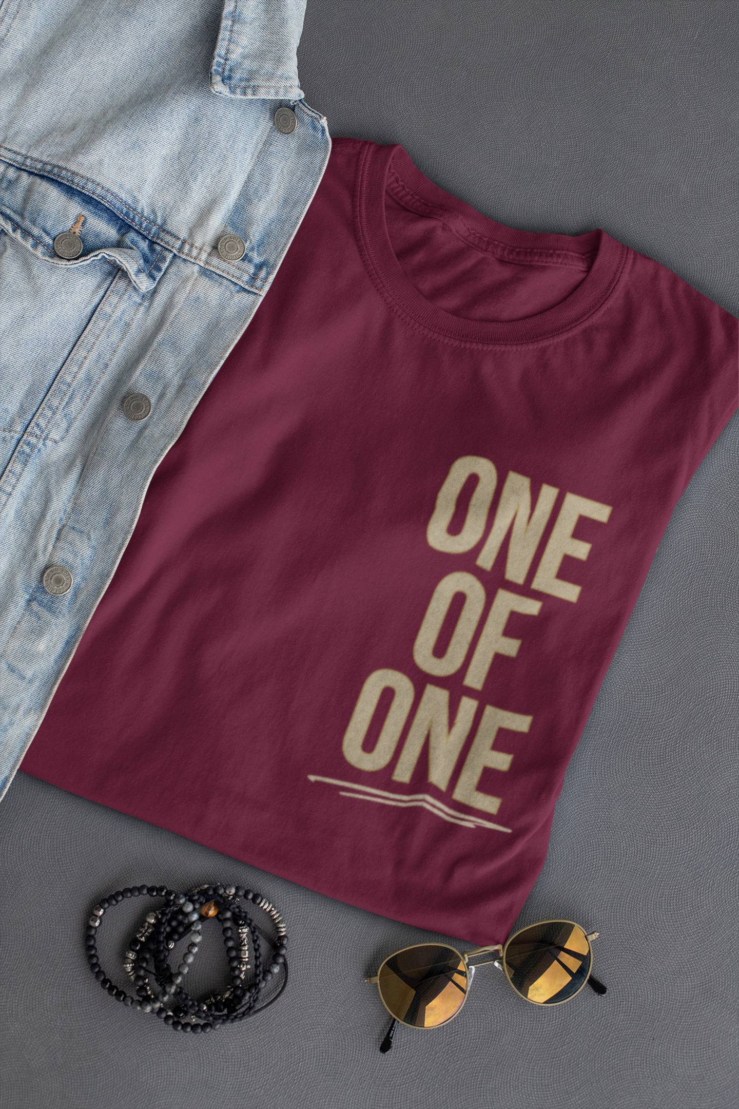 A maroon Bible verse t-shirt with 'ONE OF ONE' printed in beige block letters, styled in a flat lay with a light blue denim jacket, black beaded bracelets, and gold-framed sunglasses on a dark gray background.