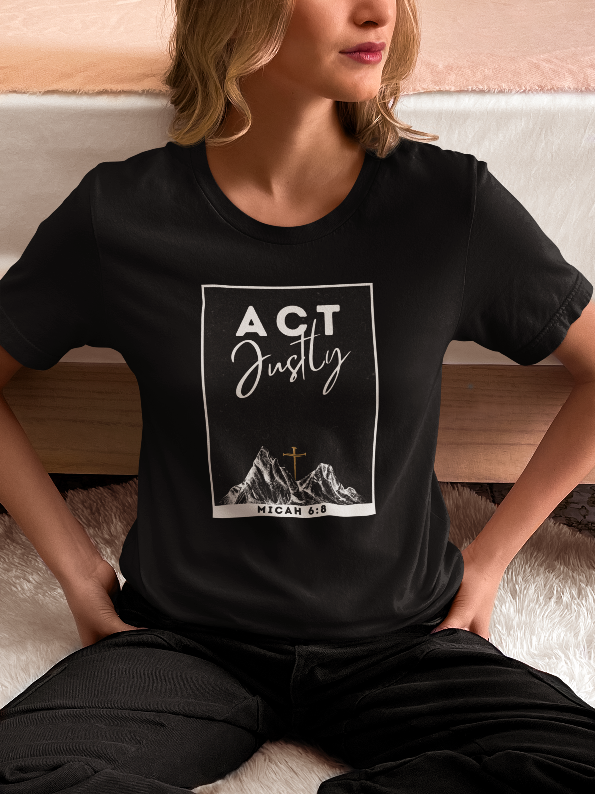 A woman wearing a black Christian Bible verse t-shirt with a white graphic print that reads 'ACT Justly' and 'MICAH 6:8' above a mountain and cross illustration, sitting on a bed.