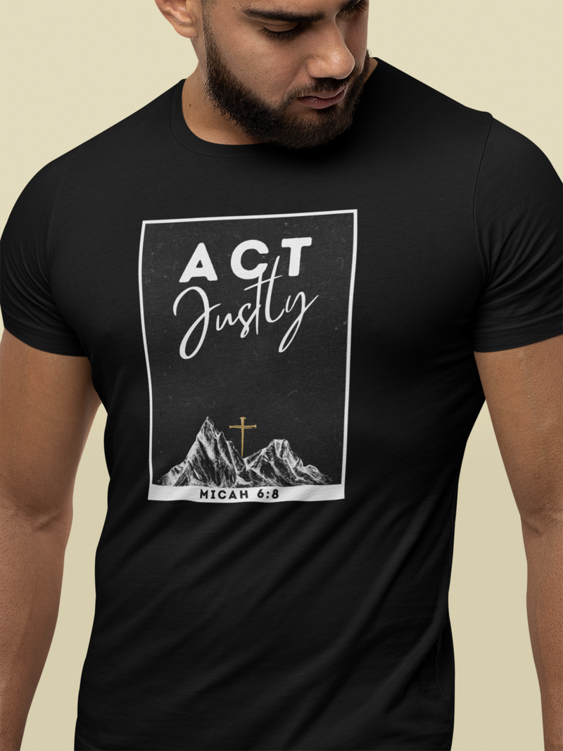 A man wearing a black Bible verse t-shirt with a white graphic print that reads 'ACT Justly' above an illustration of mountains and a cross, with 'MICAH 6:8' at the bottom, against a light beige background.