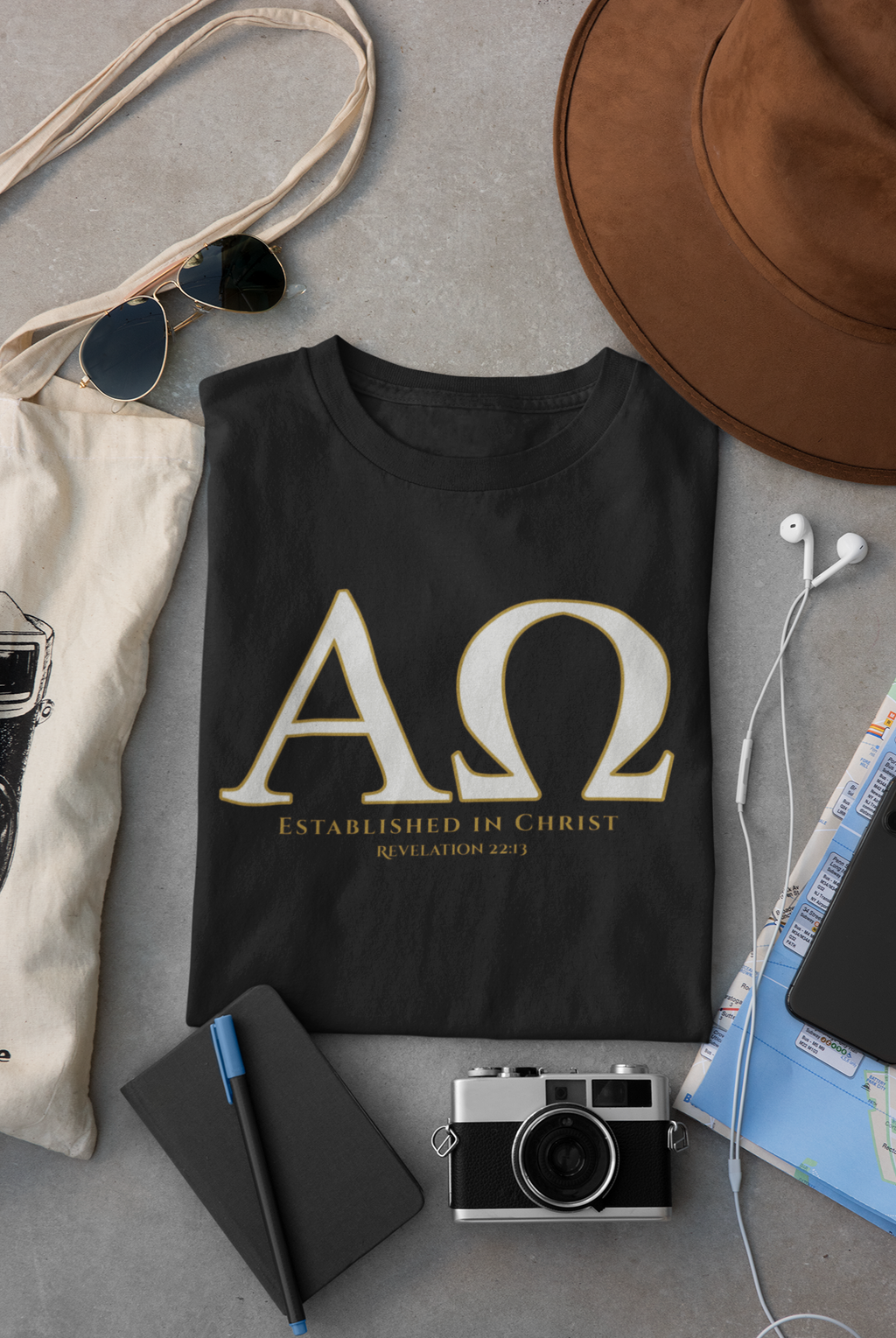 A black Christian graphic t-shirt with a gold Greek letter 'ΑΩ' and the text 'ESTABLISHED IN CHRIST REVELATION 22:13' is displayed in a styled flat-lay arrangement on a gray surface, surrounded by a brown hat, sunglasses, a canvas tote bag, a notebook, a camera, and earphones.