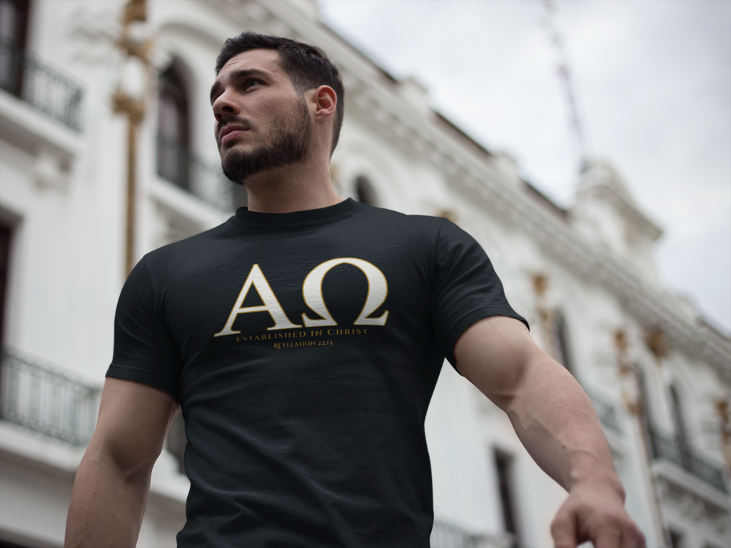 A man wearing a black Christian conversation starter t-shirt with a large gold Greek letter 'ΑΩ' and the text 'ESTABLISHED IN CHRIST' REVELATION 22:13' printed on the chest, set against a blurred background of a white building.