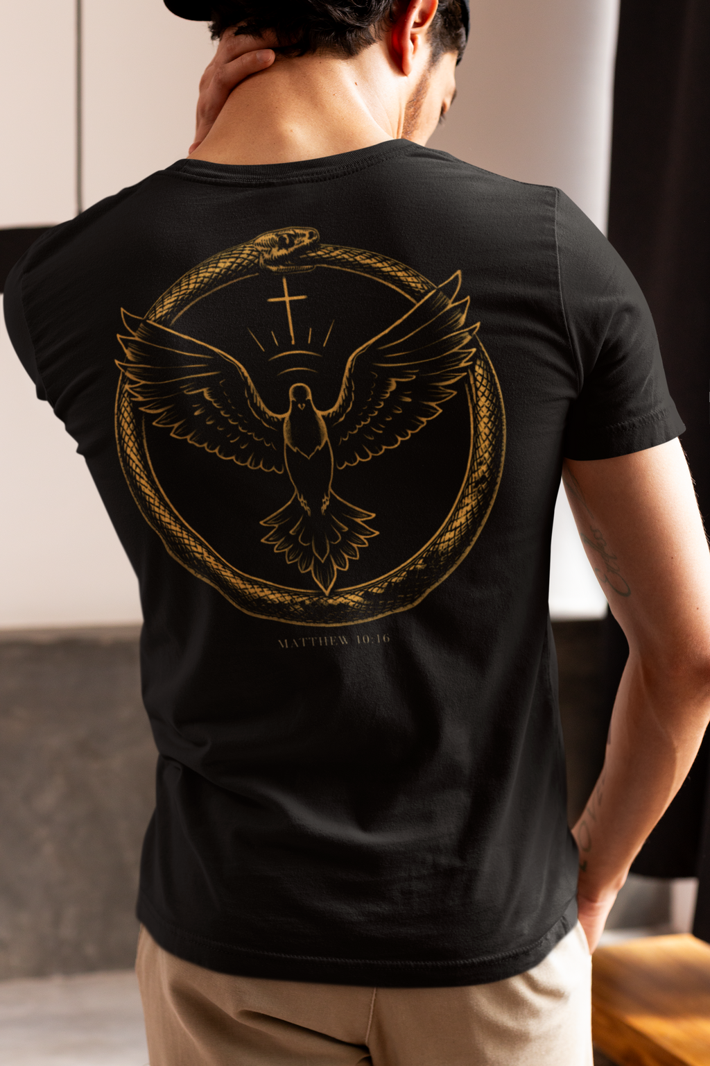 A man shown from behind wearing a black Christian graphic t-shirt with a large gold-colored graphic of a dove with wings spread inside a circular frame, with the text 'Matthew 10:16' printed below. The background is a blurred indoor setting.