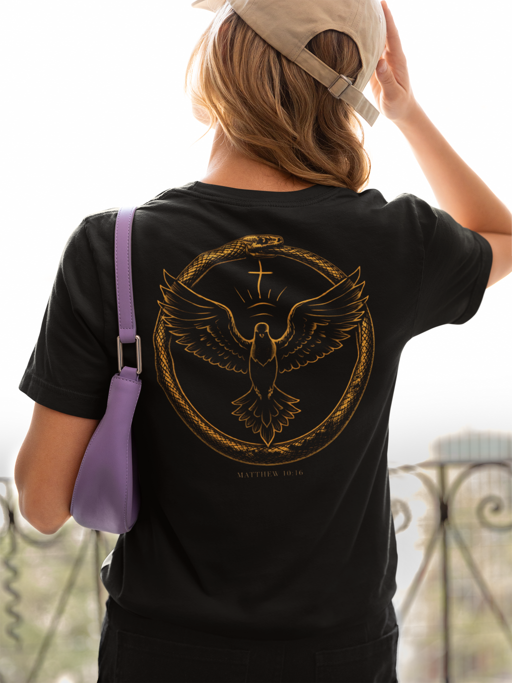 Person wearing a black faith based t-shirt with a gold dove encircled by a snake with the text 'Matthew 10:16' design on the back, holding a beige cap.