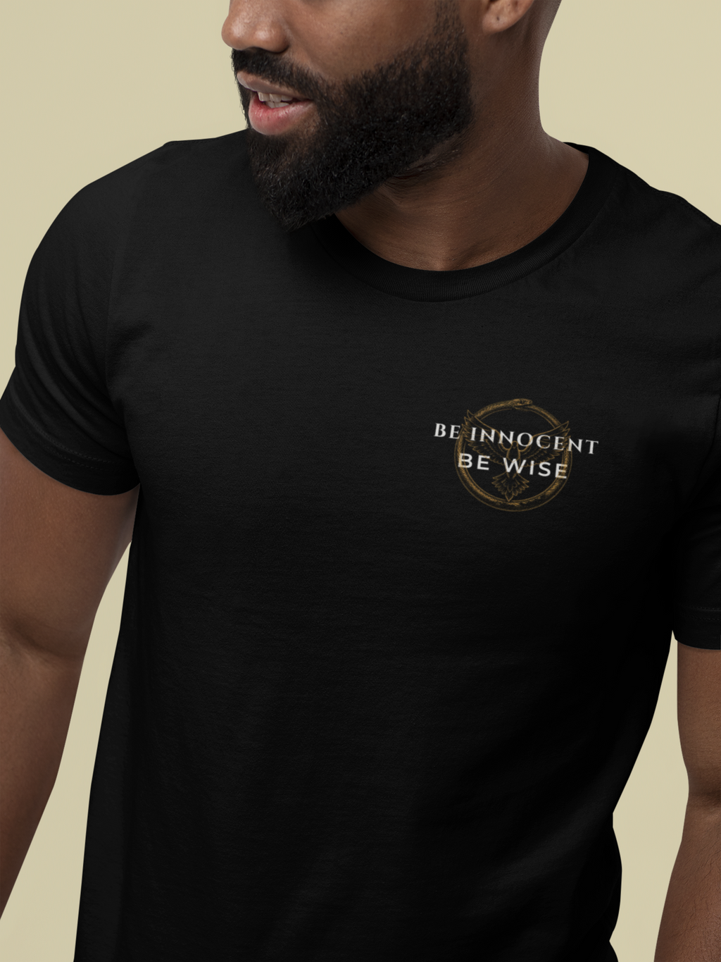 A man wearing a black Christian Bible verse t-shirt with a gold circular emblem and the text 'BE INNOCENT BE WISE' printed on the left chest, set against a solid beige background.
