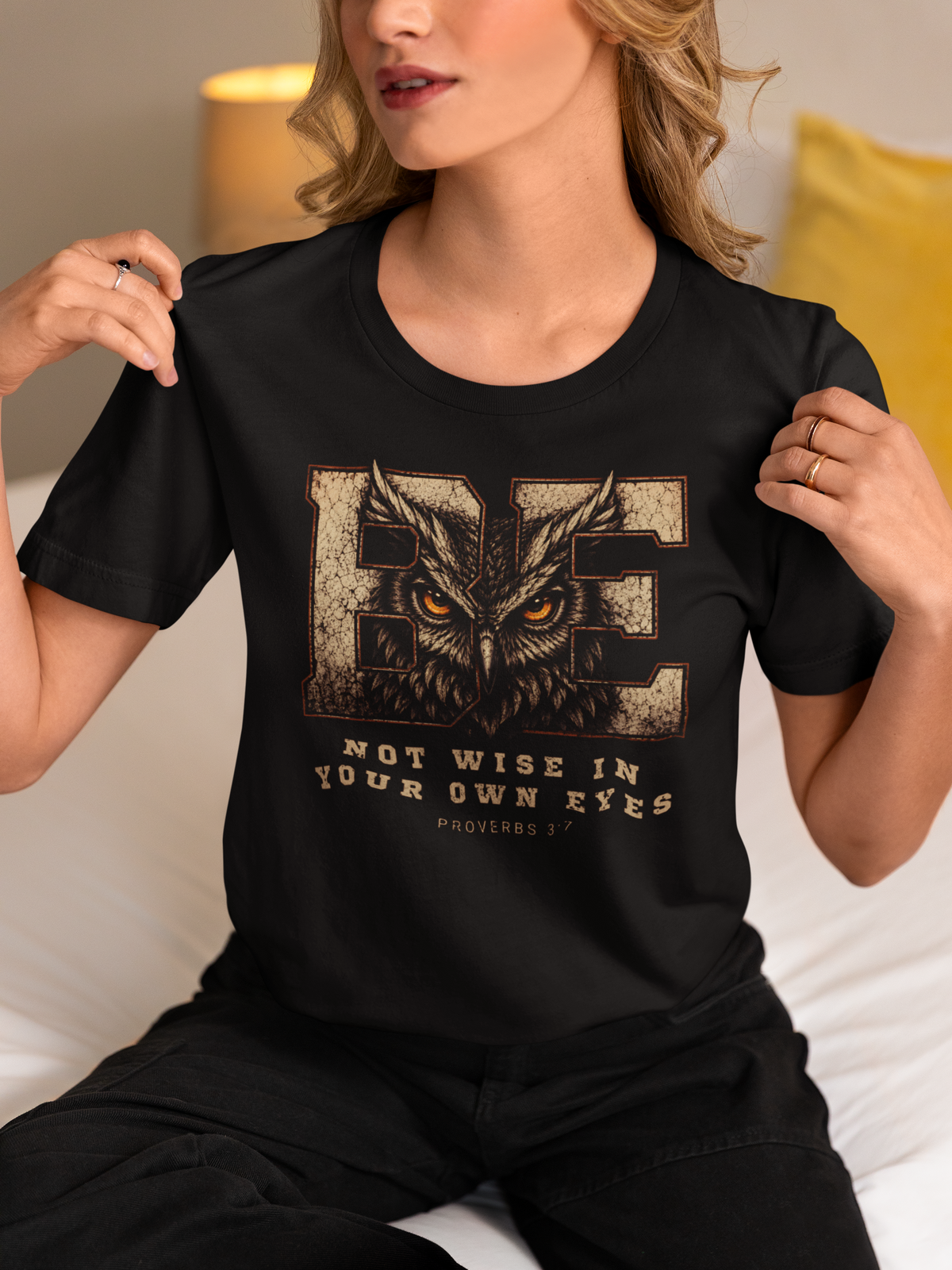 A woman wearing a black short-sleeve Christian graphic T-shirt with a large owl design and the text 'BE NOT WISE IN YOUR OWN EYES' from Proverbs 3:7, posed in a cozy indoor setting.