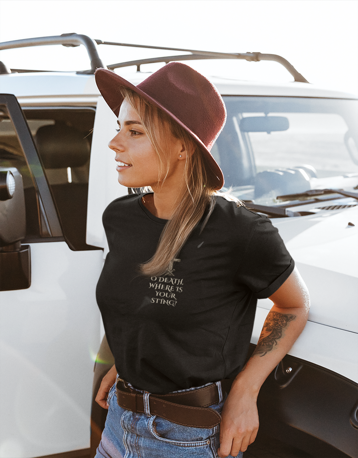 A woman wearing a black Christian graphic t-shirt and a brown wide-brim hat leans against a white vehicle. The t-shirt features the text 'O DEADL WHERE IS YOUR STRING' in white.