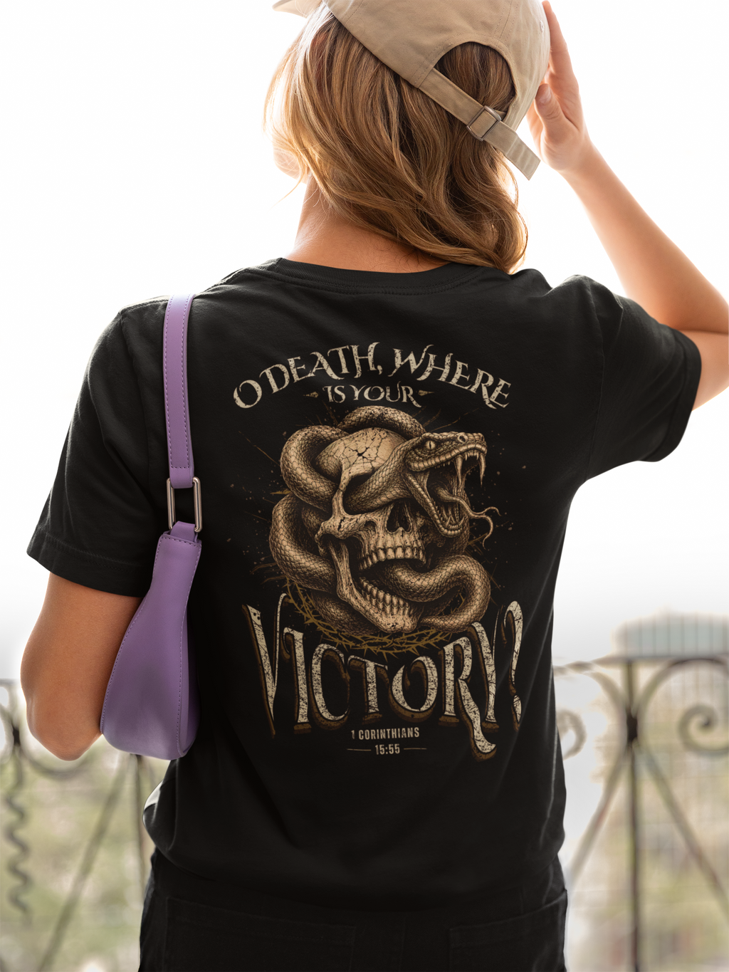 A woman viewed from behind wearing a black Christian graphic T-shirt with a large snake-and-skull design and the text 'O DEATH, WHERE IS YOUR VICTORY!'. She is also wearing a beige cap and carrying a purple shoulder bag, standing outdoors near a railing.