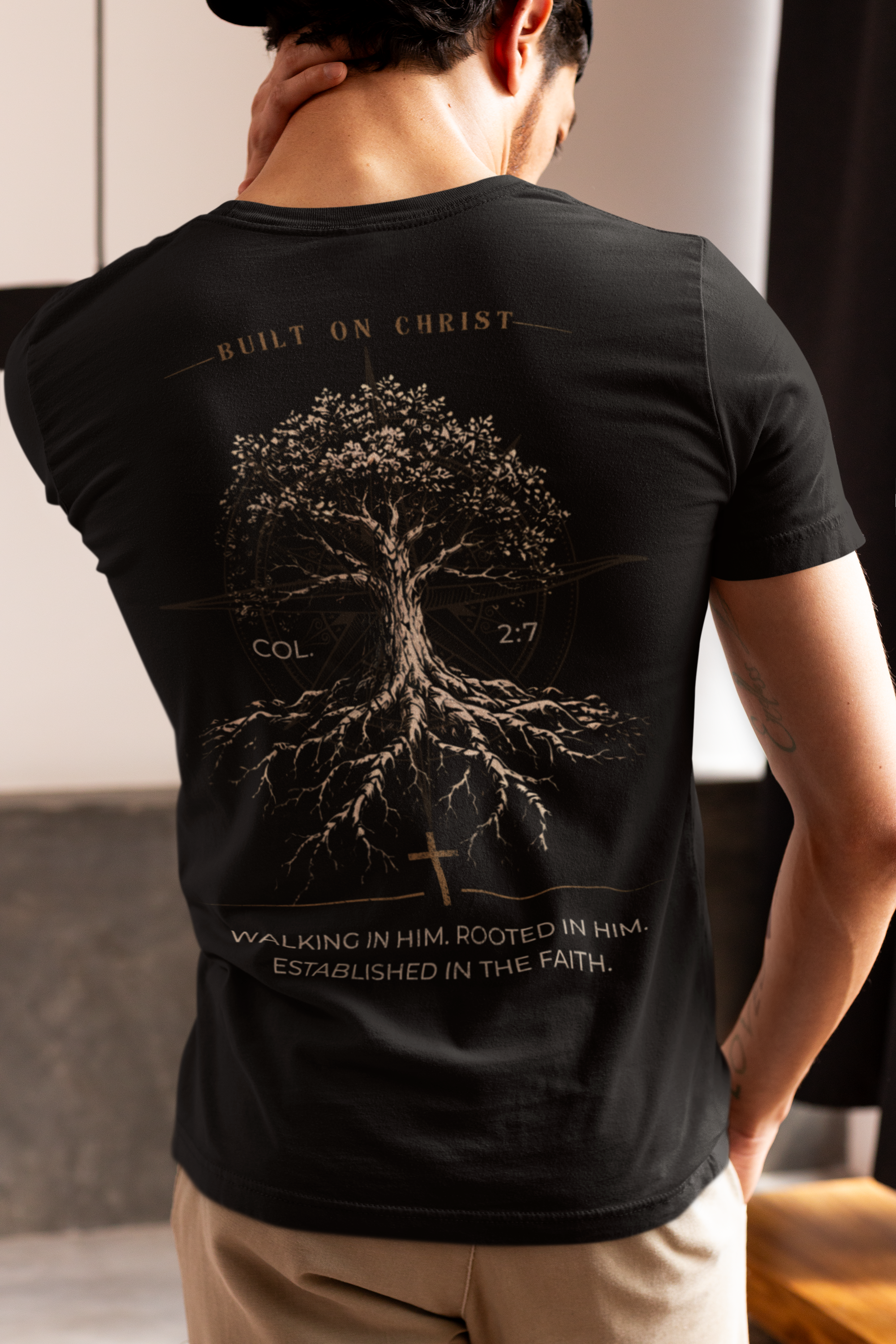 A man viewed from behind wearing a black Christian graphic t-shirt with a large graphic print on the back. The print features a tree of life with roots, the text 'BUILT ON CHRIST' at the top and 'WALKING IN HIM, ROOTED IN HIM, ESTABLISHED IN THE FAITH' at the bottom, and 'COL 2:7' on either side of the tree.