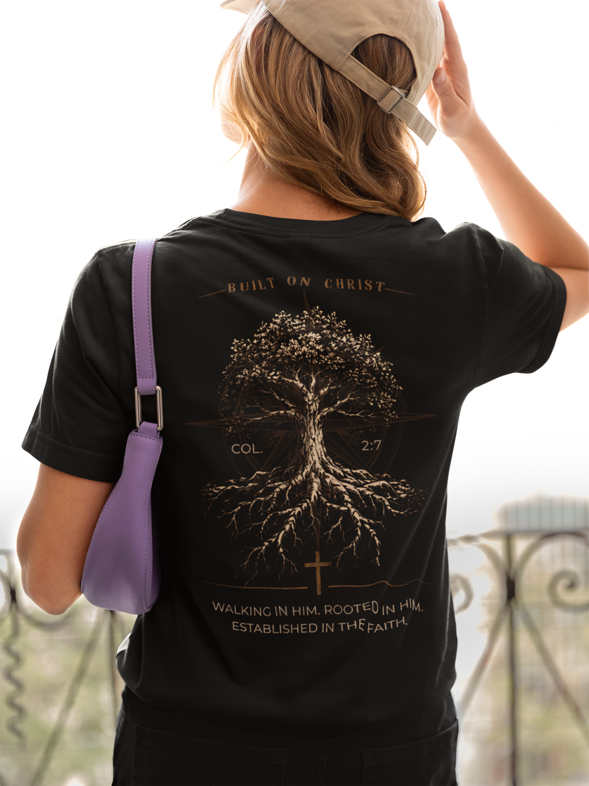 A woman viewed from behind wearing a black Christian graphic t-shirt with a large gold graphic of a tree of life and religious text on the back, along with a light beige baseball cap and a purple shoulder bag.