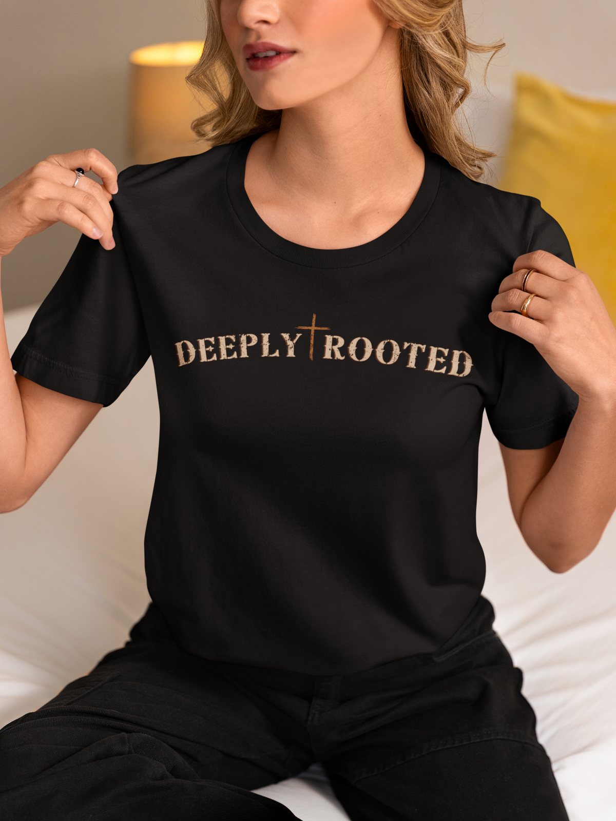 A woman wearing a black short-sleeve Christian graphic t-shirt with 'DEEPLY ROOTED' printed in a metallic gold textured font across the chest, posed in a cozy indoor setting.