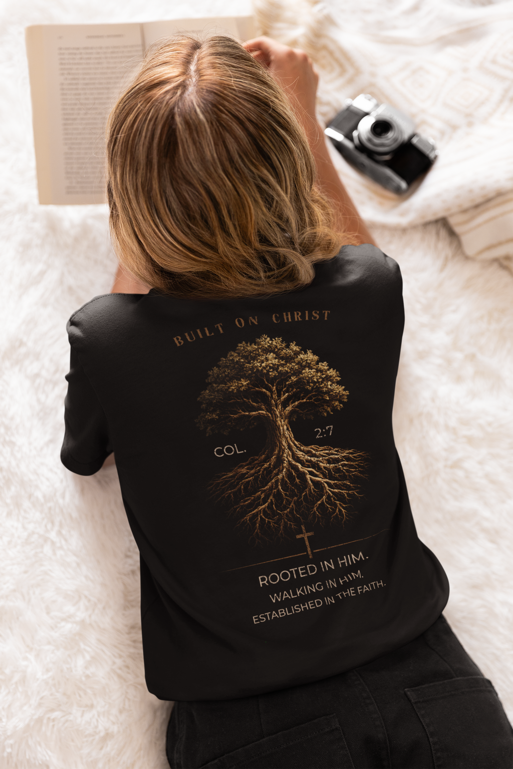A woman lying on a white faux-fur surface, viewed from behind, wearing a black Christian Bible verse T-shirt with a large gold tree graphic and Bible verse text on the back. A book, camera, and blanket are visible around her.