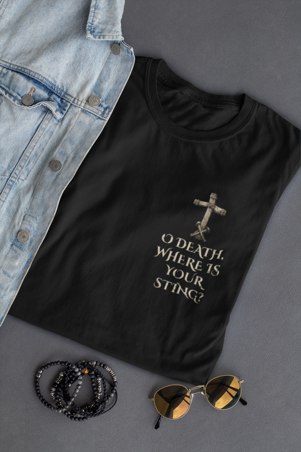 A black Chrisitan graphic t-shirt with a distressed cross and the text 'O DEATH WHERE IS YOUR STING?' in a light beige font, styled in a flat lay with a denim jacket, beaded bracelets, and sunglasses on a gray background.
