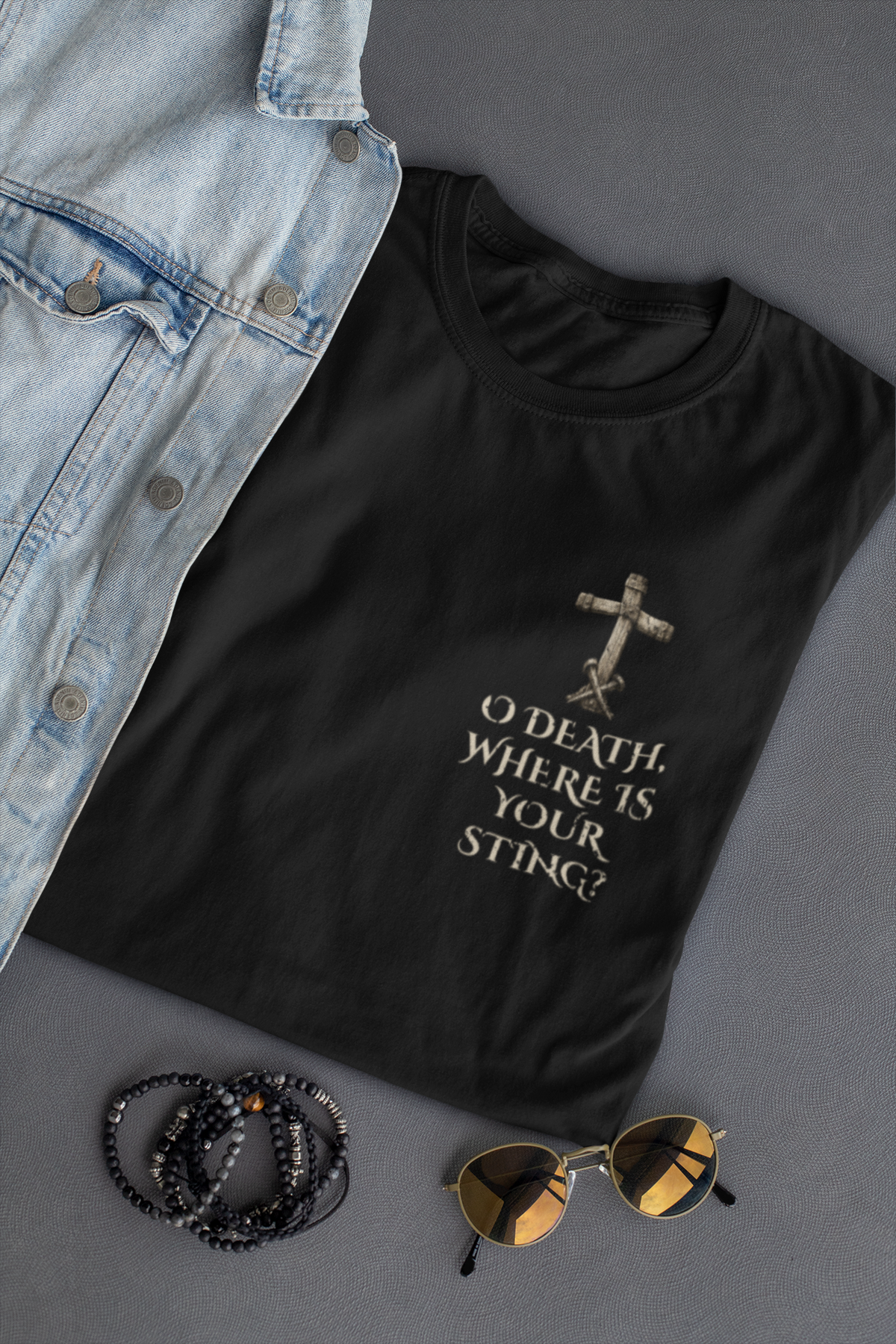 A black Chrisitan graphic t-shirt with a distressed cross and the text 'O DEATH WHERE IS YOUR STING?' in a light beige font, styled in a flat lay with a denim jacket, beaded bracelets, and sunglasses on a gray background.