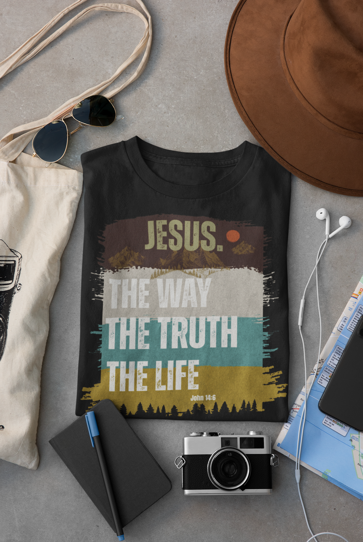 A black Christian graphic T-shirt laid flat on a gray surface, surrounded by lifestyle props including a canvas tote bag, sunglasses, a brown hat, earphones, a notebook, a camera, and a phone. The shirt features a distressed multicolor print with the text 'JESUS. The way, the truth , the life."