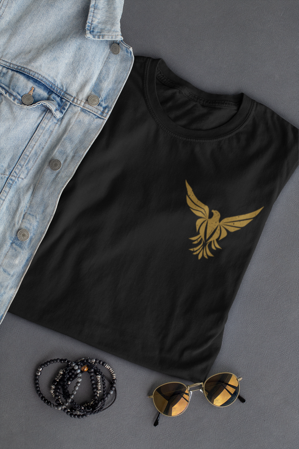 A black short-sleeve  Christian faith T-shirt with a gold bird logo on the left chest, styled in a flat lay with a light blue denim shirt, black beaded bracelets, and gold-framed sunglasses on a gray background.