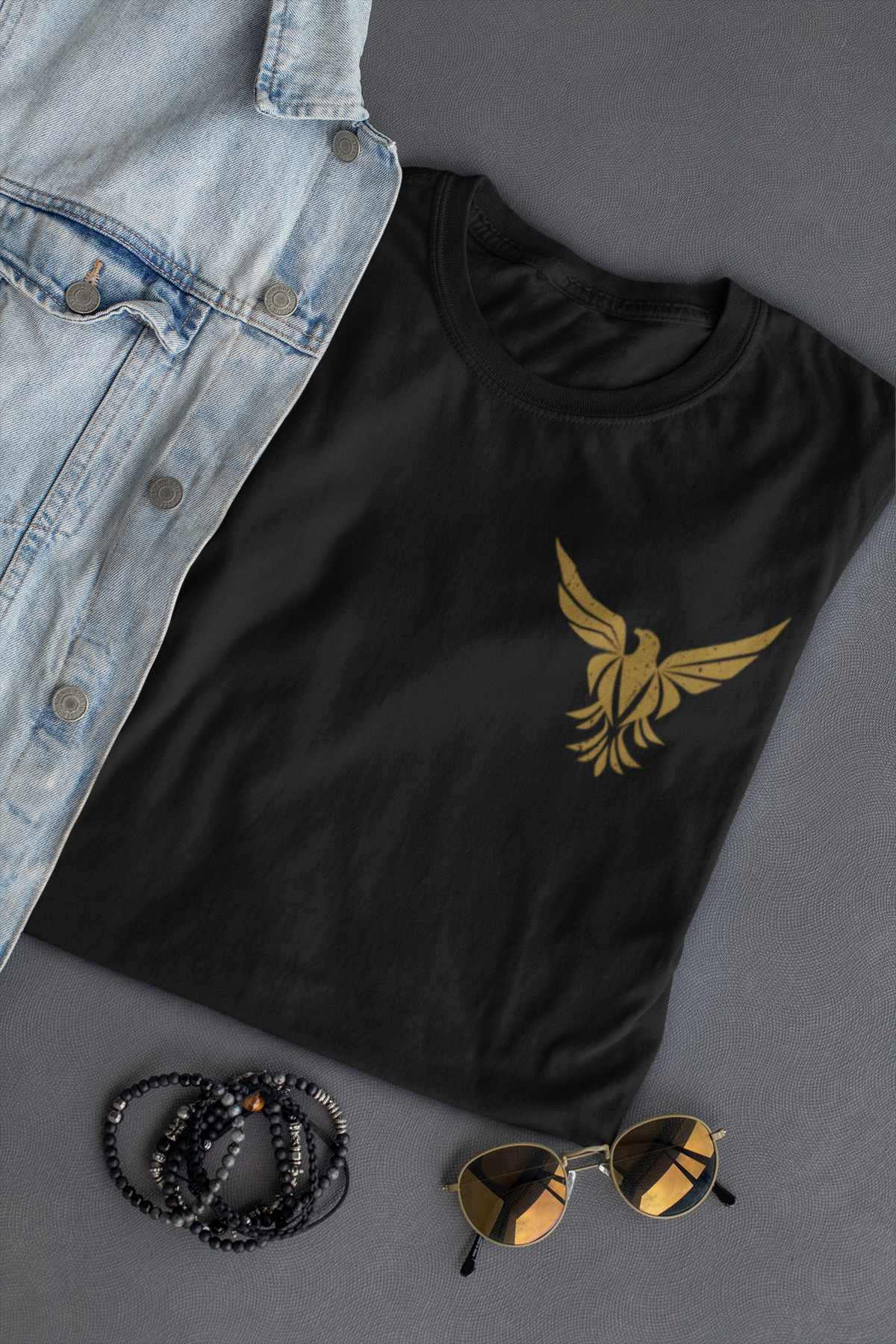 A black short-sleeve  Christian faith T-shirt with a gold bird logo on the left chest, styled in a flat lay with a light blue denim shirt, black beaded bracelets, and gold-framed sunglasses on a gray background.