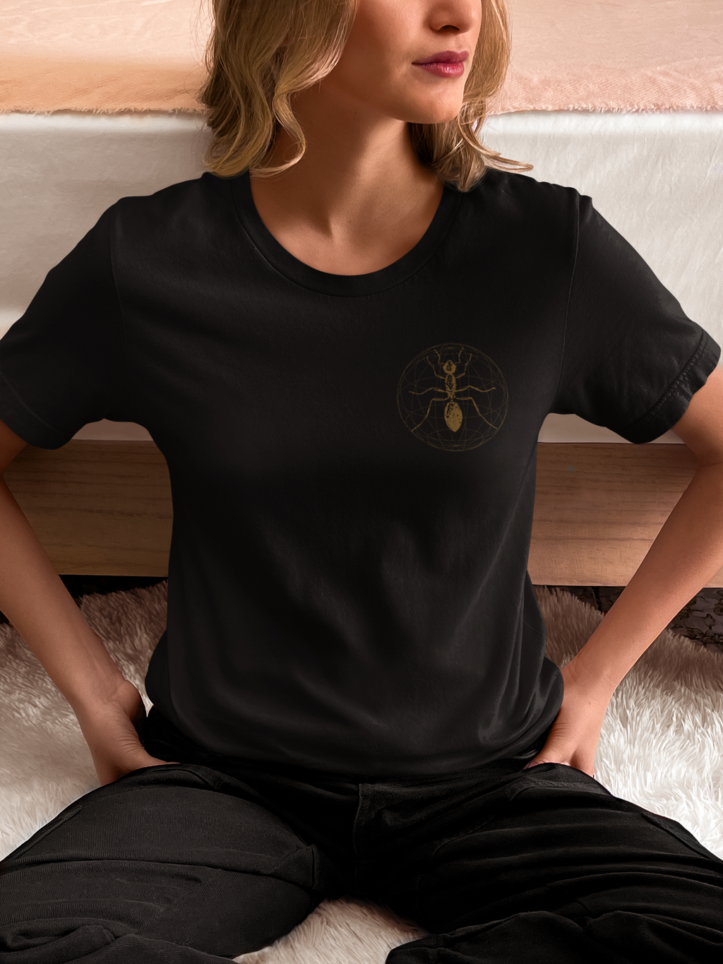 A woman wearing a black faith t-shirt with a small gold ant graphic on the left chest, sitting on a white rug in front of a wooden headboard.
