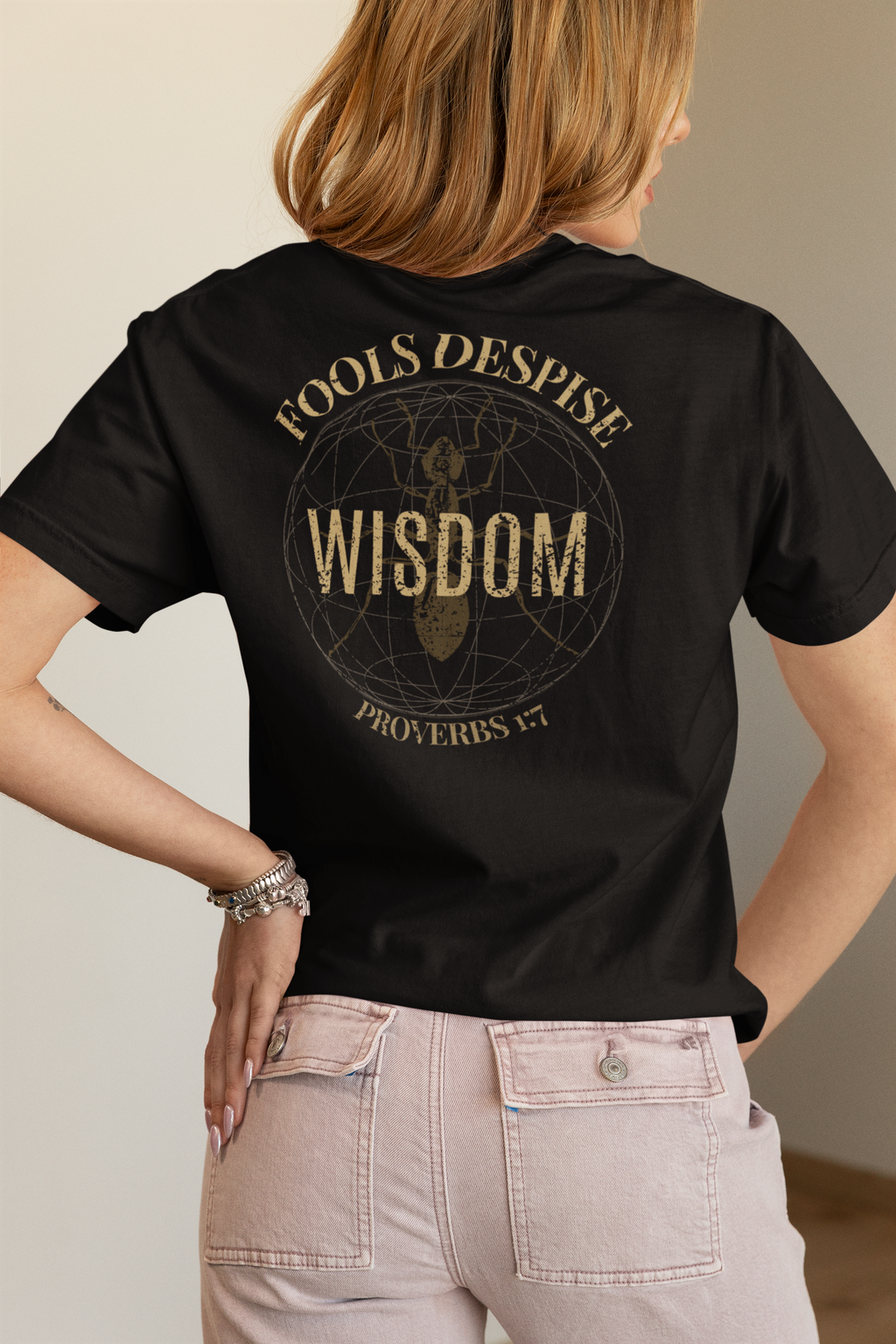 A woman shown from the back wearing a black bible verse T-shirt with a large distressed gold graphic on the back that reads “FOOLS DESPISE WISDOM PROVERBS 1:7” around a circular emblem, paired with light beige pants.