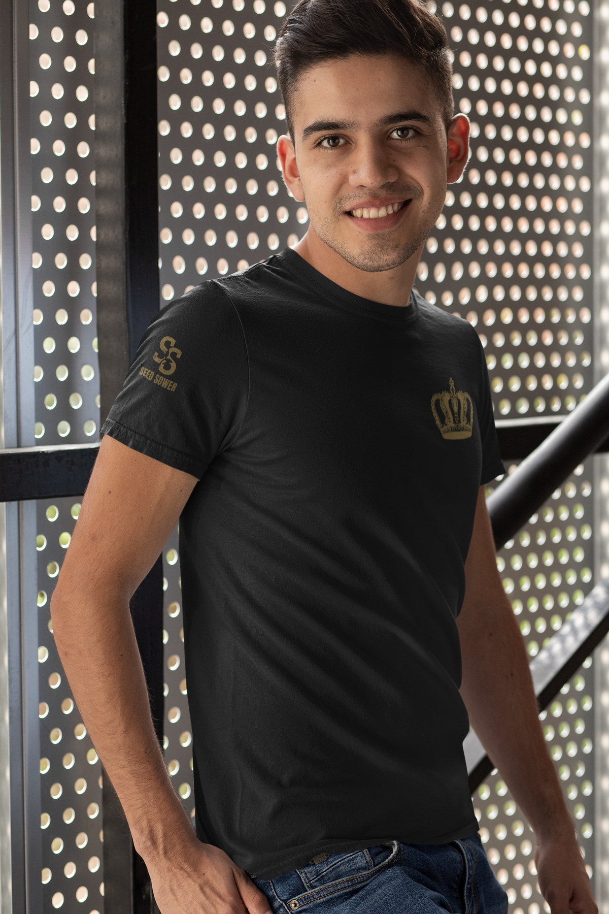 A smiling man wearing a black short-sleeve Christian graphic t-shirt with a gold crown logo on the left chest and a gold sleeve logo, standing in front of a perforated metal screen.