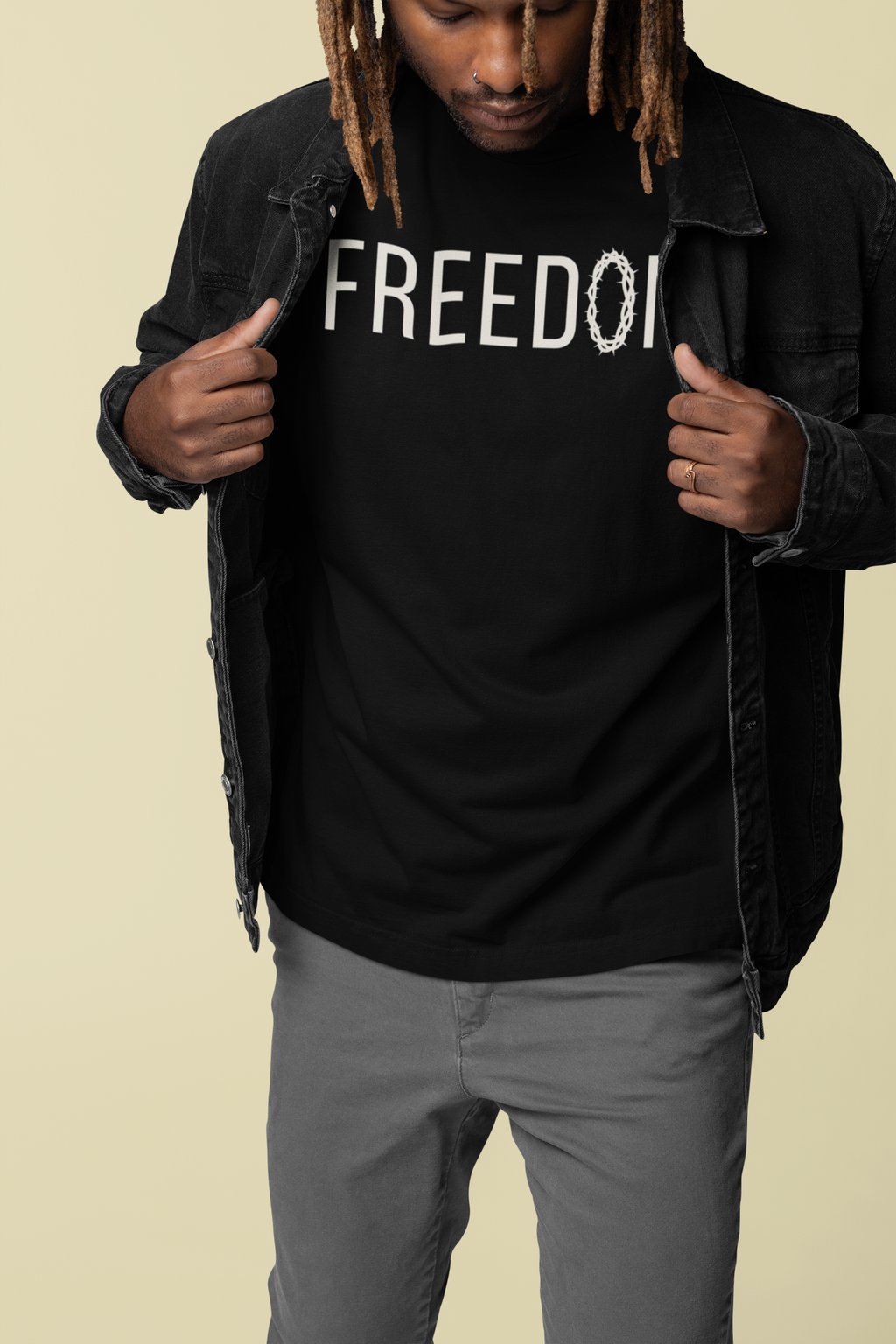 A man wearing a black Christian faith t-shirt with 'FREEDOM' printed in white across the chest, layered under a dark denim jacket, paired with gray pants, set against a solid light beige background.