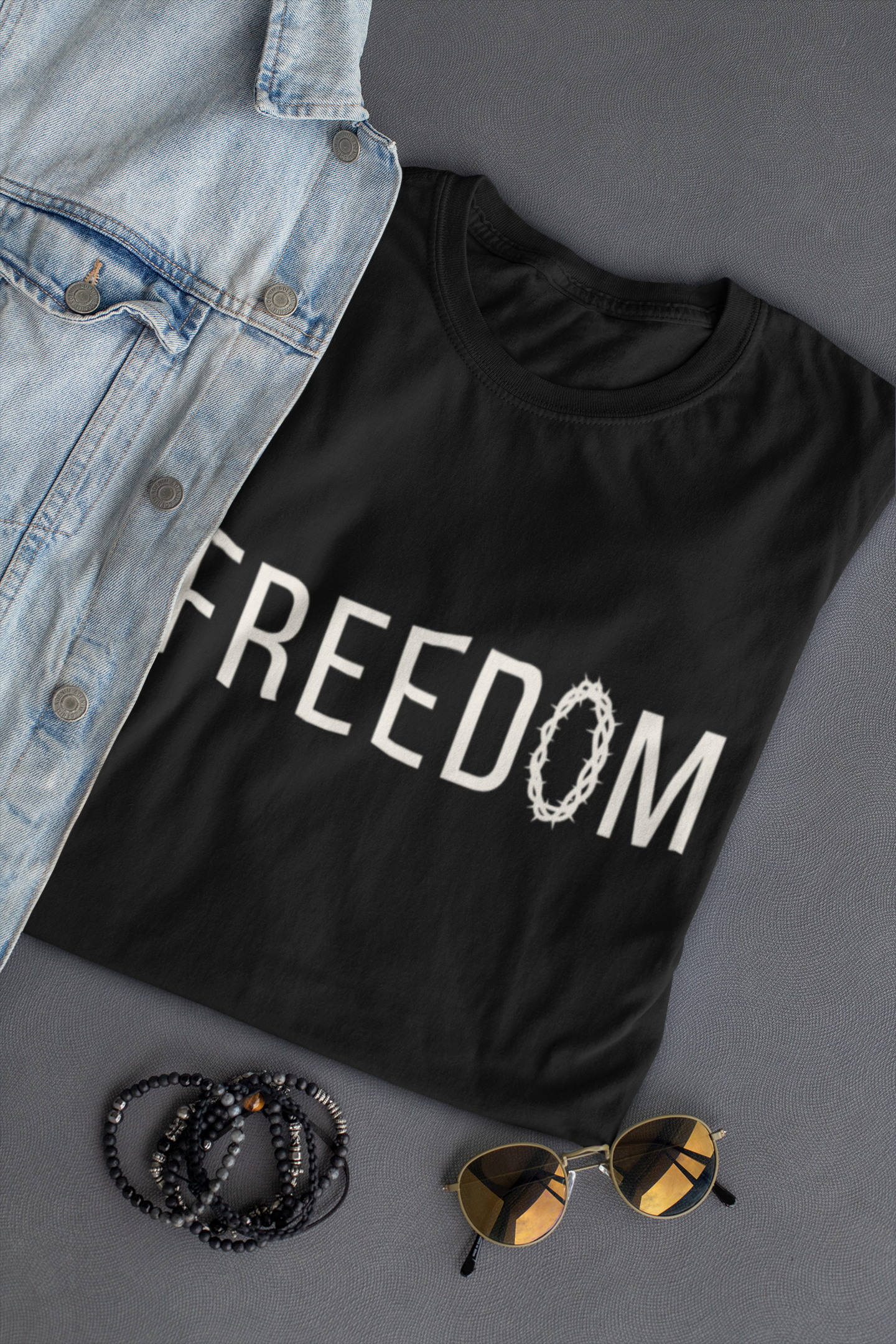 A black Chrisitan t-shirt with 'FREEDOM' printed in white across the chest, styled in a flat-lay with a light blue denim jacket, black beaded bracelets, and gold-framed sunglasses on a dark gray background.