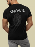 A man wearing a black Christian Bible verse t-shirt from the back, featuring the word 'KNOWN.' in white text above a gray fingerprint graphic made of script text that is words of affirmation from the Bible.