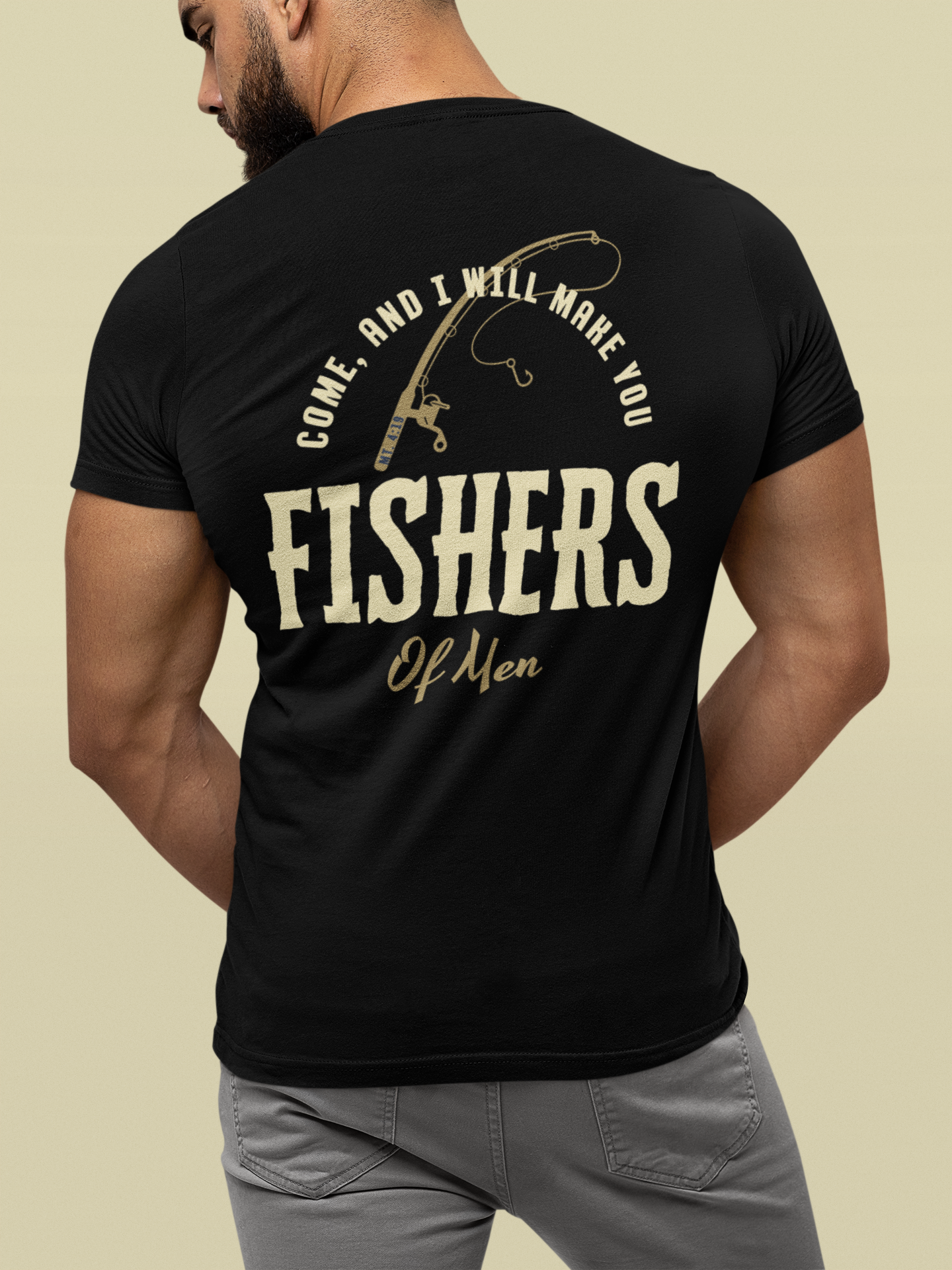 A man shown from the back wearing a black faith based t-shirt with a large cream-colored graphic on the back that reads 'COME, AND I WILL MAKE YOU FISHERS OF MEN' with a fishing rod illustration, paired with gray pants against a solid beige background.