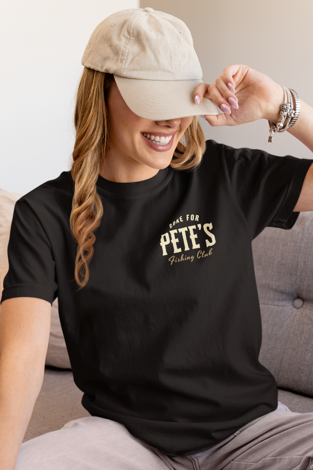 A woman wearing a black Bible verse t-shirt with a cream graphic that reads 'GONE FOR PETE'S Fishing Club' and a beige baseball cap, sitting on a gray couch.