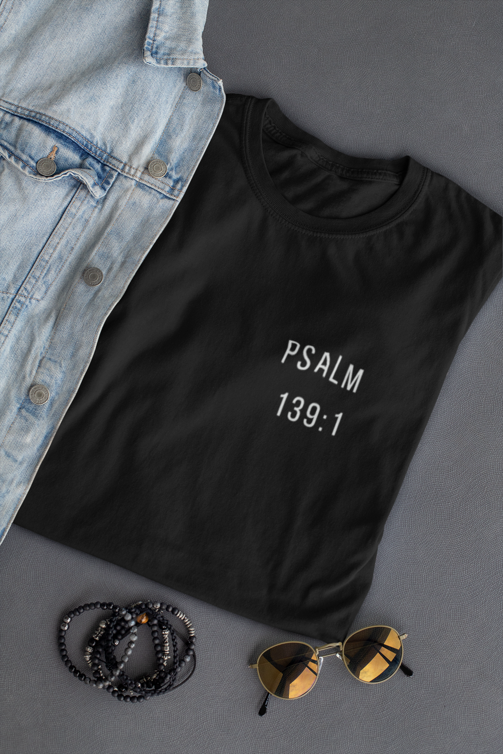 A black short-sleeve Christian T-shirt with 'PSALM 139:1' printed in white text on the chest, styled in a flat lay with a light blue denim jacket, black beaded bracelets, and gold-framed sunglasses on a dark gray background.
