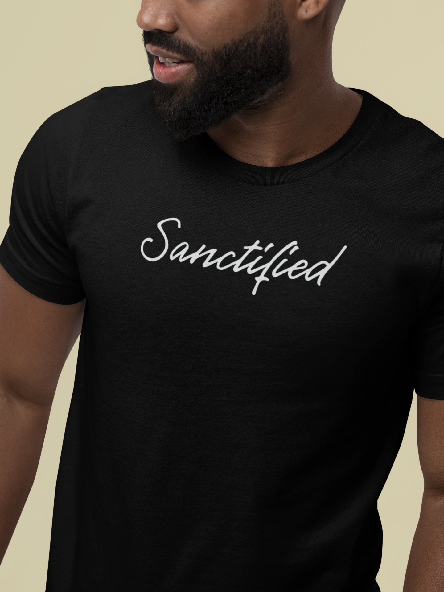 A close-up of a man wearing a black Christian t-shirt with the word 'Sanctified' printed in white script across the chest, set against a solid beige background.