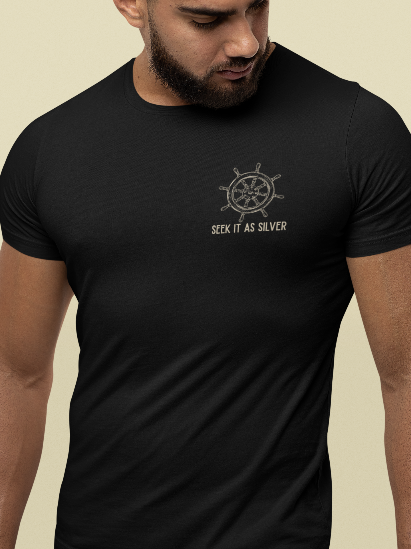 A man wearing a black Christian t-shirt with a silver ship's wheel graphic and the text 'SEEK IT AS SILVER' printed on the chest, set against a solid beige background.