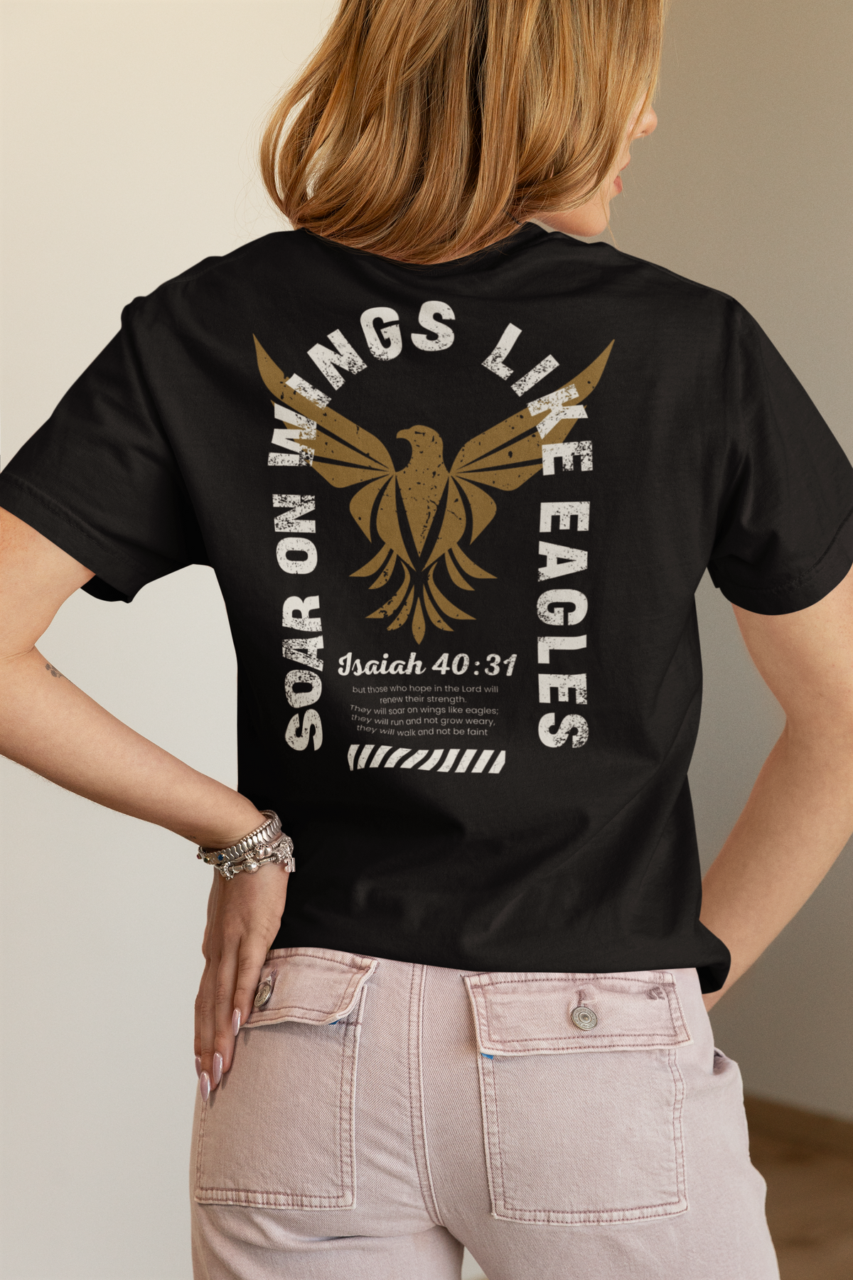 A woman shown from the back wearing a black Christian faith short-sleeve T-shirt with a large gold eagle graphic and white text reading 'Soar on wings like eagles' and 'Isaiah 40:31'. She is also wearing light pink jeans against a neutral indoor background.