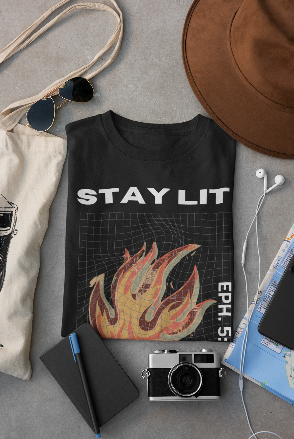 A black Christian graphic T-shirt laid flat on a gray surface, surrounded by lifestyle props including a brown hat, sunglasses, tote bag, earphones, notebook, camera, and map. The shirt has large white text reading 'STAY LIT' across the chest a fiery orange/yellow graphic below, and vertical white text on the right side with the verse Ephesians 5:8.