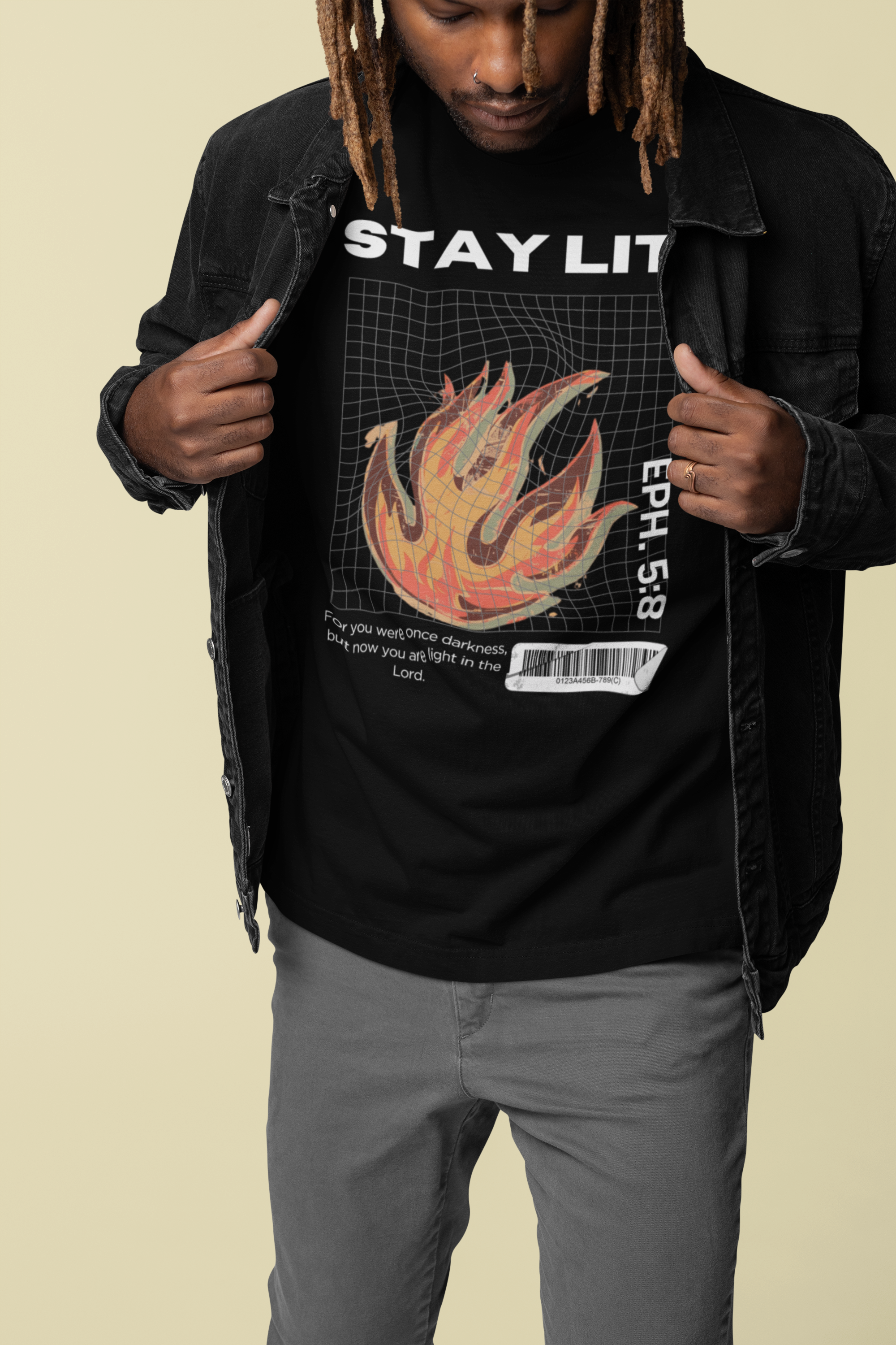 A man wearing a black Christian graphic t-shirt with a flame design and the text 'STAY LIT' and 'EP. 5:8', layered under an open black denim jacket, paired with gray pants, against a solid beige background.