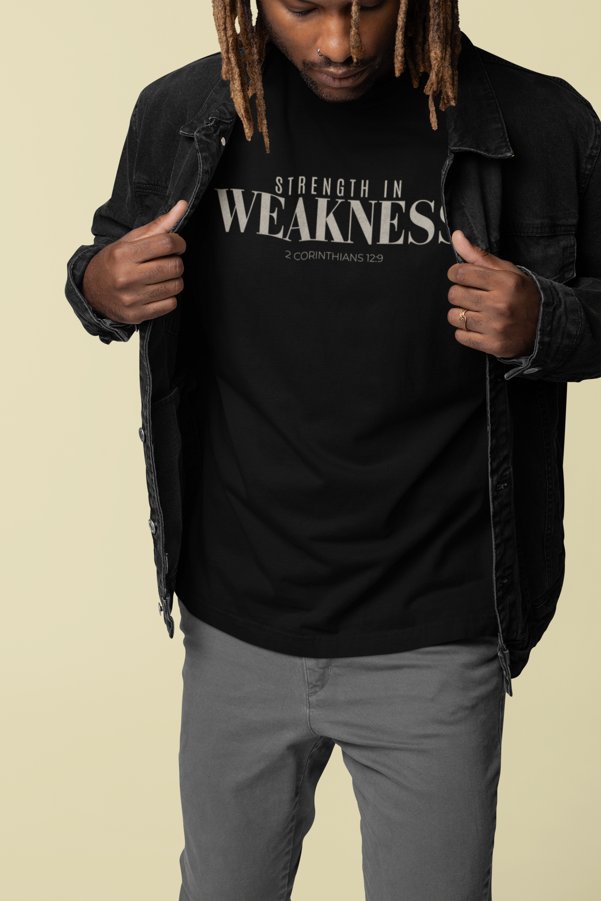A man wearing a black Christian graphic t-shirt with the text 'STRENGTH IN WEAKNESS 2 CORINTHIANS 12:9' in white, styled with a black denim jacket and gray pants against a solid beige background.