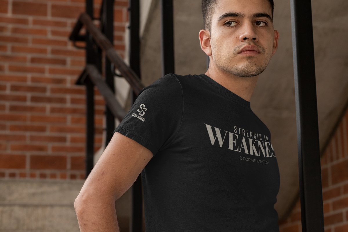 A man wearing a black Christianity t-shirt with 'STRENGTH IN WEAKNES' printed in white on the chest, posed in front of a brick wall and staircase.