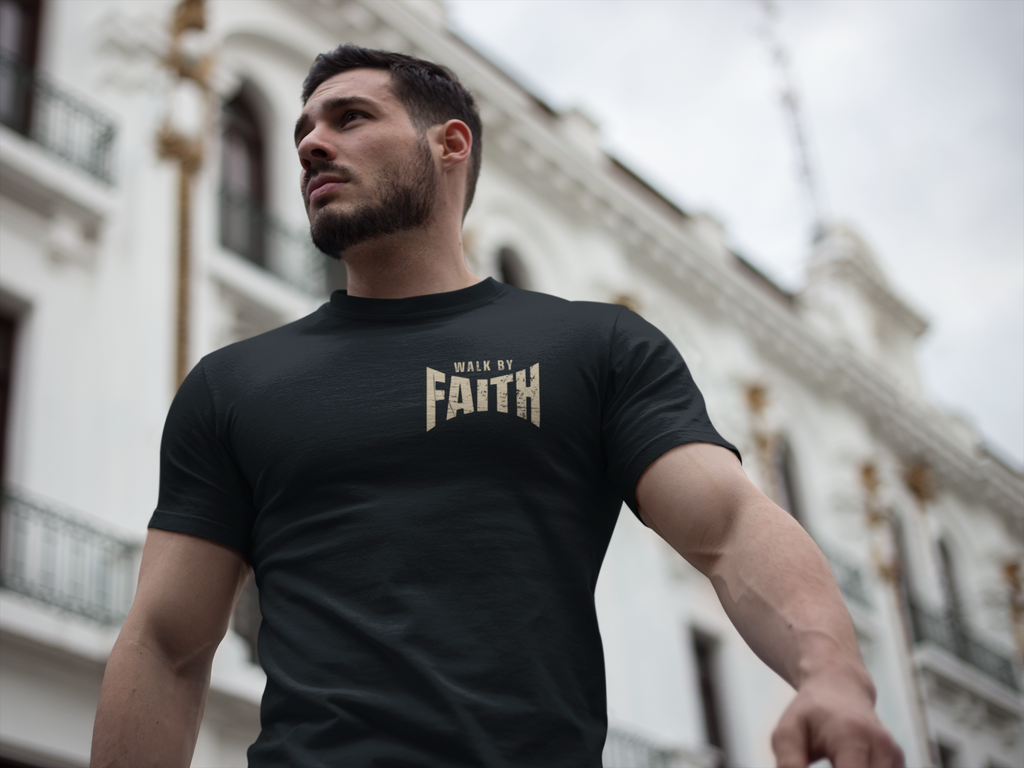 A muscular man wearing a black Christian graphic t-shirt with 'WALK BY FAITH' printed in gold on the chest, photographed from a low angle with a blurred white building in the background.