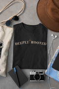 A black Christian graphic t-shirt with 'DEEPLY ROOTED' printed in beige across the chest, styled in a flat lay with a brown hat, sunglasses, canvas tote bag, notebook, camera, and earphones on a gray surface.