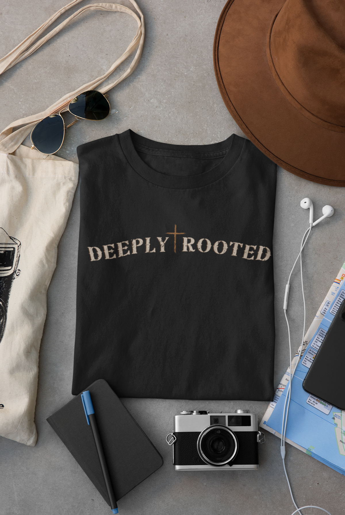A black Christian graphic t-shirt with 'DEEPLY ROOTED' printed in beige across the chest, styled in a flat lay with a brown hat, sunglasses, canvas tote bag, notebook, camera, and earphones on a gray surface.