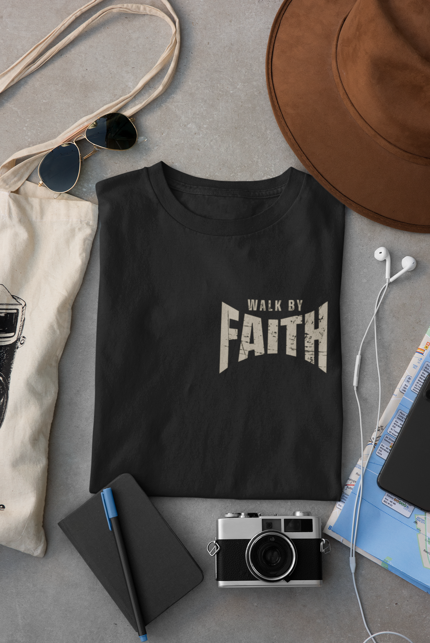 A black crewneck Christian graphic t-shirt with a distressed white graphic reading 'WALK BY FAITH' is displayed in a styled flat-lay arrangement on a gray surface, surrounded by a brown hat, sunglasses, a canvas tote bag, a notebook, earphones, and a camera.