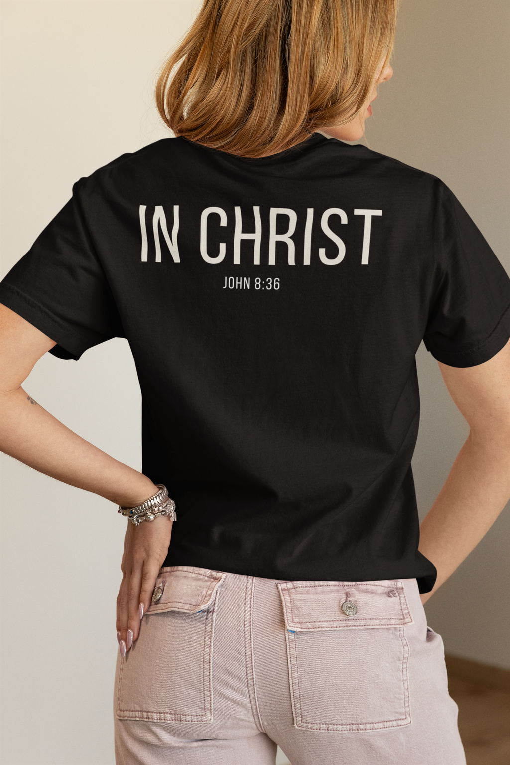 A woman shown from the back wearing a black Christian conversation starter T-shirt with the words 'IN CHRIST' and 'JOHN 8:36' printed in white across the upper back, paired with light pink pants.