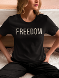 A woman wearing a black faith apparel t-shirt with the word 'FREEDOM' printed in white across the chest, sitting on a bed with her hands on her hips.